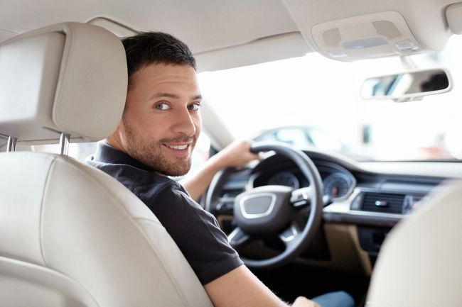Un uomo sorridente in un'auto guarda indietro dal posto di guida, con interni beige e volante visibile.