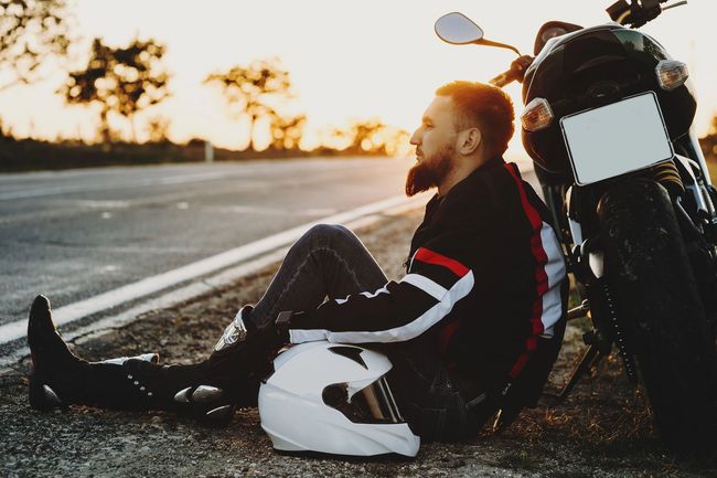 Un motociclista seduto accanto a una moto parcheggiata al tramonto sul ciglio della strada.