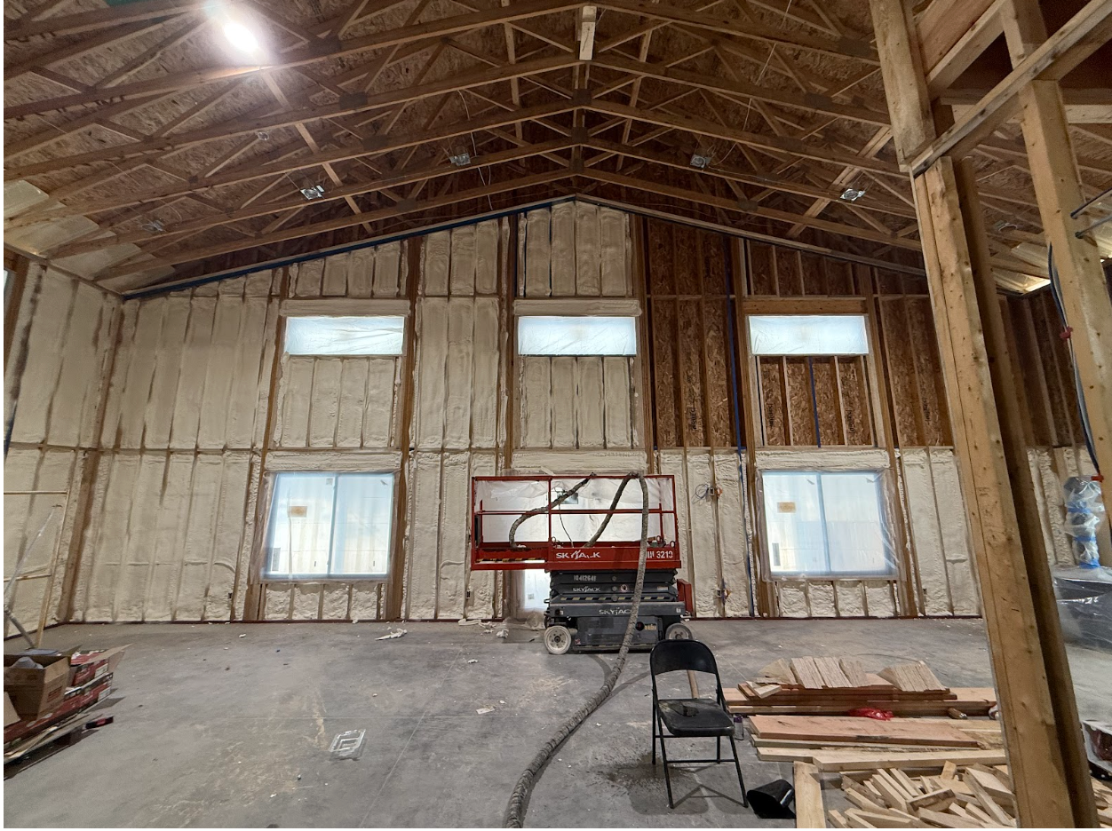 Interior view of a building under construction, spray foam insulation on walls, windows, lift equipment present.