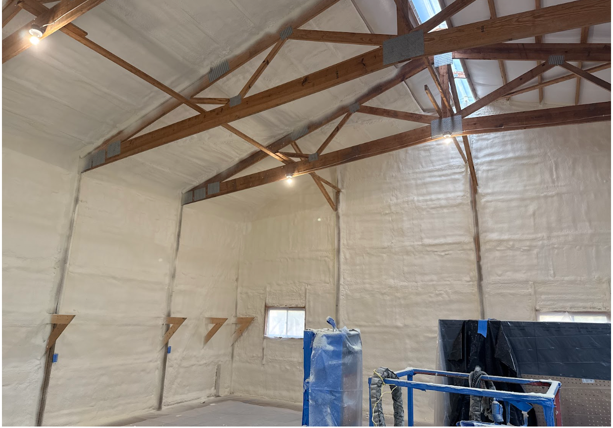 Interior of a building with white insulation on walls and ceiling, wooden beams, and a small window.
