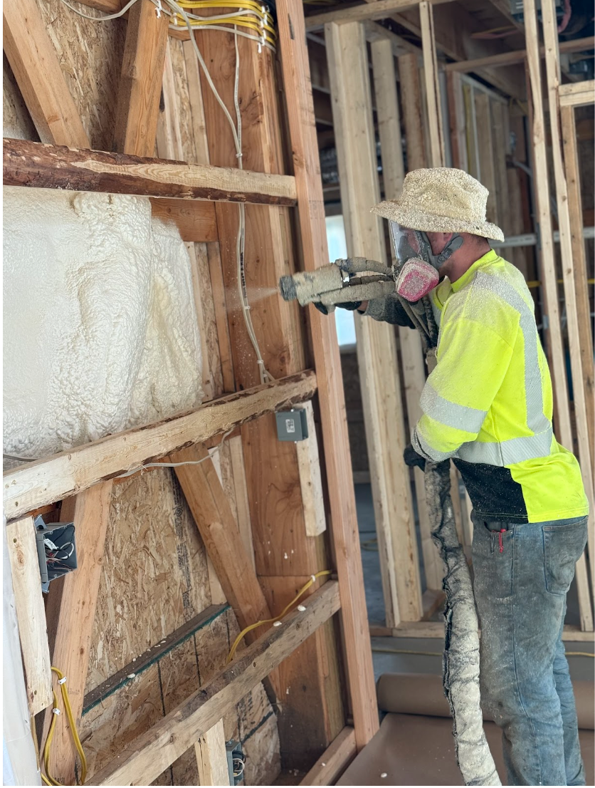 Construction worker spraying insulation foam on a wooden wall, wearing safety gear in a building.