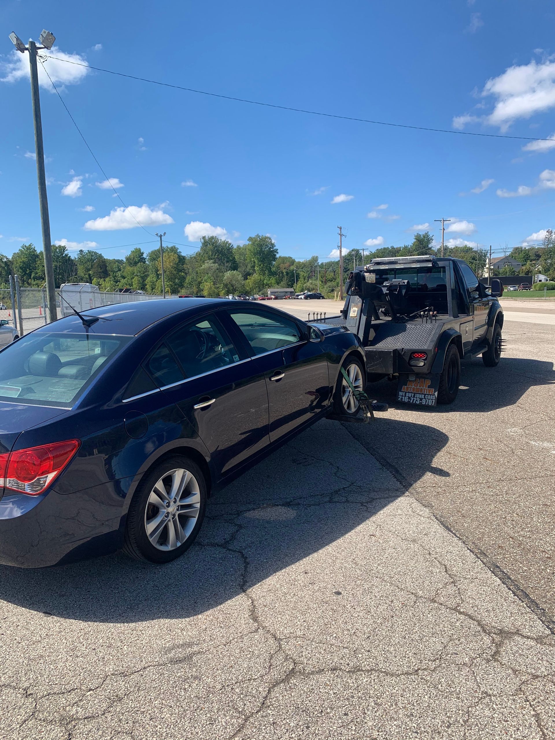 Black sedan being towed on a trailer in a sunny parking lot under a blue sky