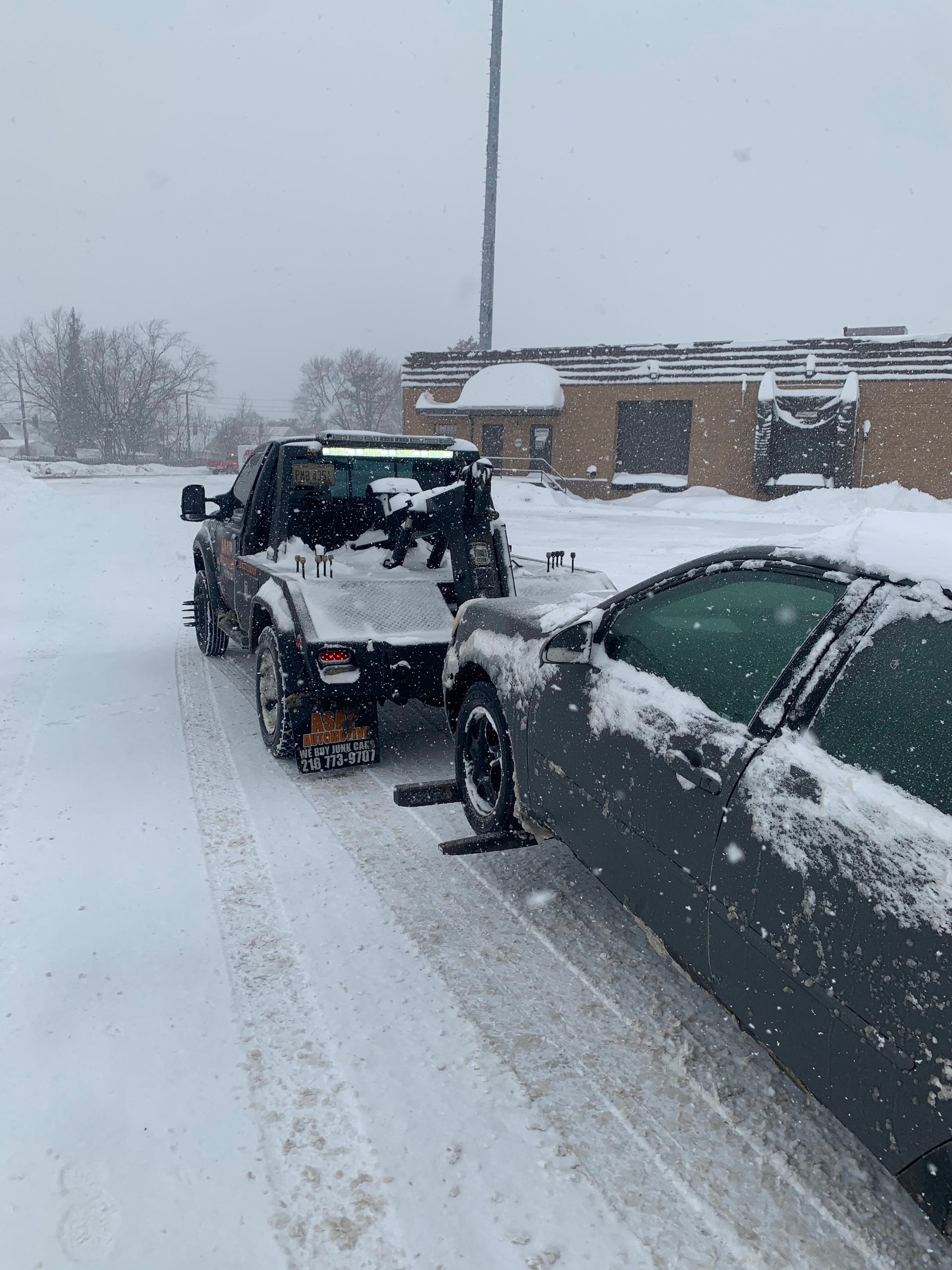 Snow-covered car towing a black off-road vehicle in a snowy parking lot.