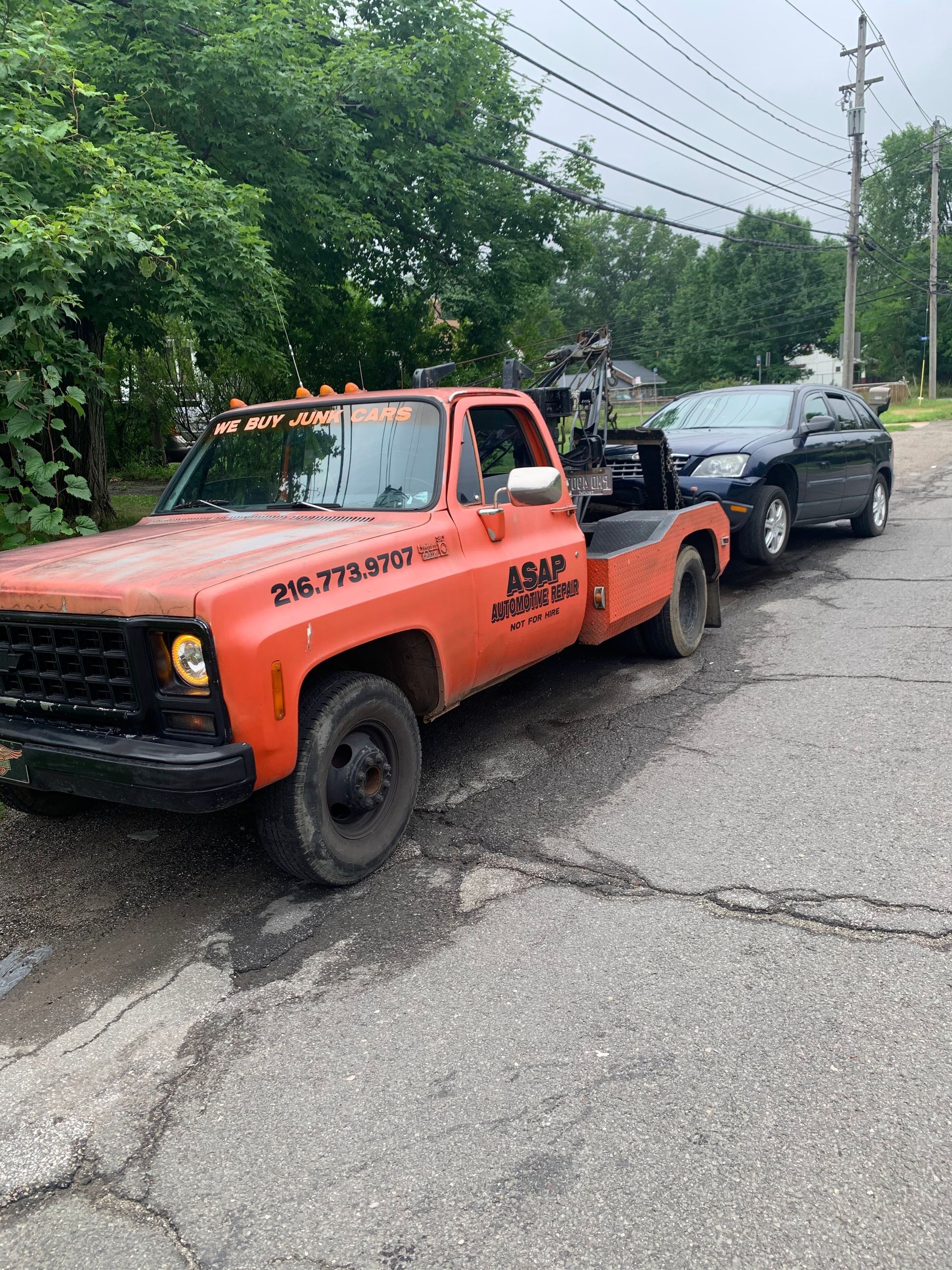 Orange tow truck parked on a roadside, with a black SUV behind it and trees along the street.