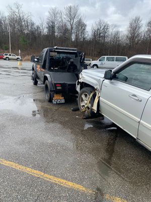 Two damaged vehicles collide on a wet roadside, with debris and tow equipment visible.