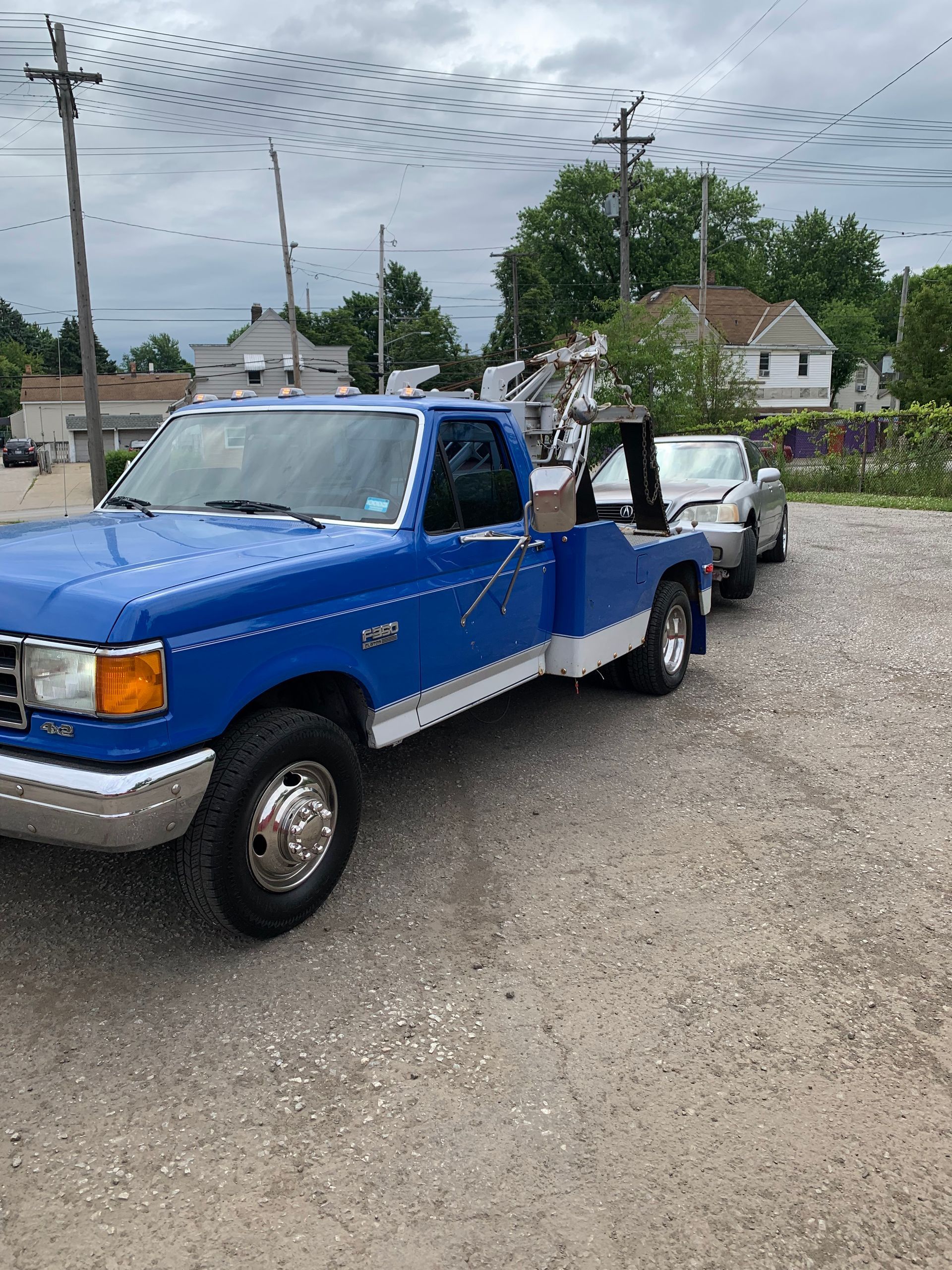 Blue pickup truck parked on gravel with a trailer loaded with potted plants in a residential area