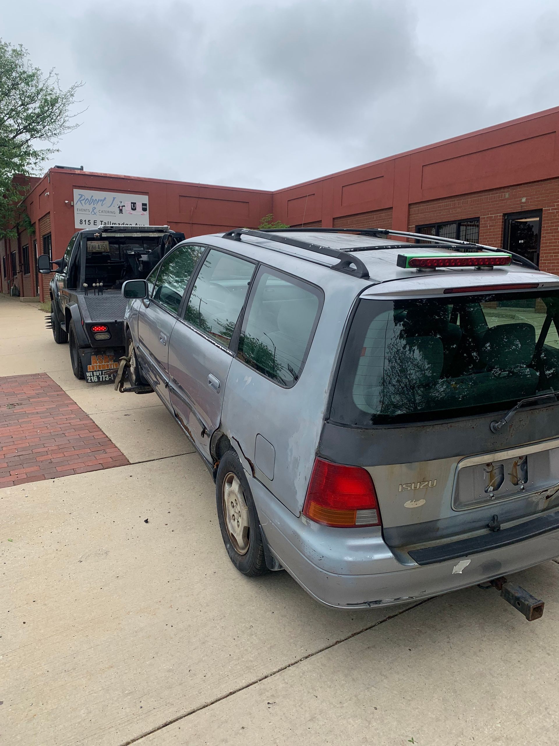 Silver minivan parked on a sidewalk beside a red brick building, with another vehicle ahead.