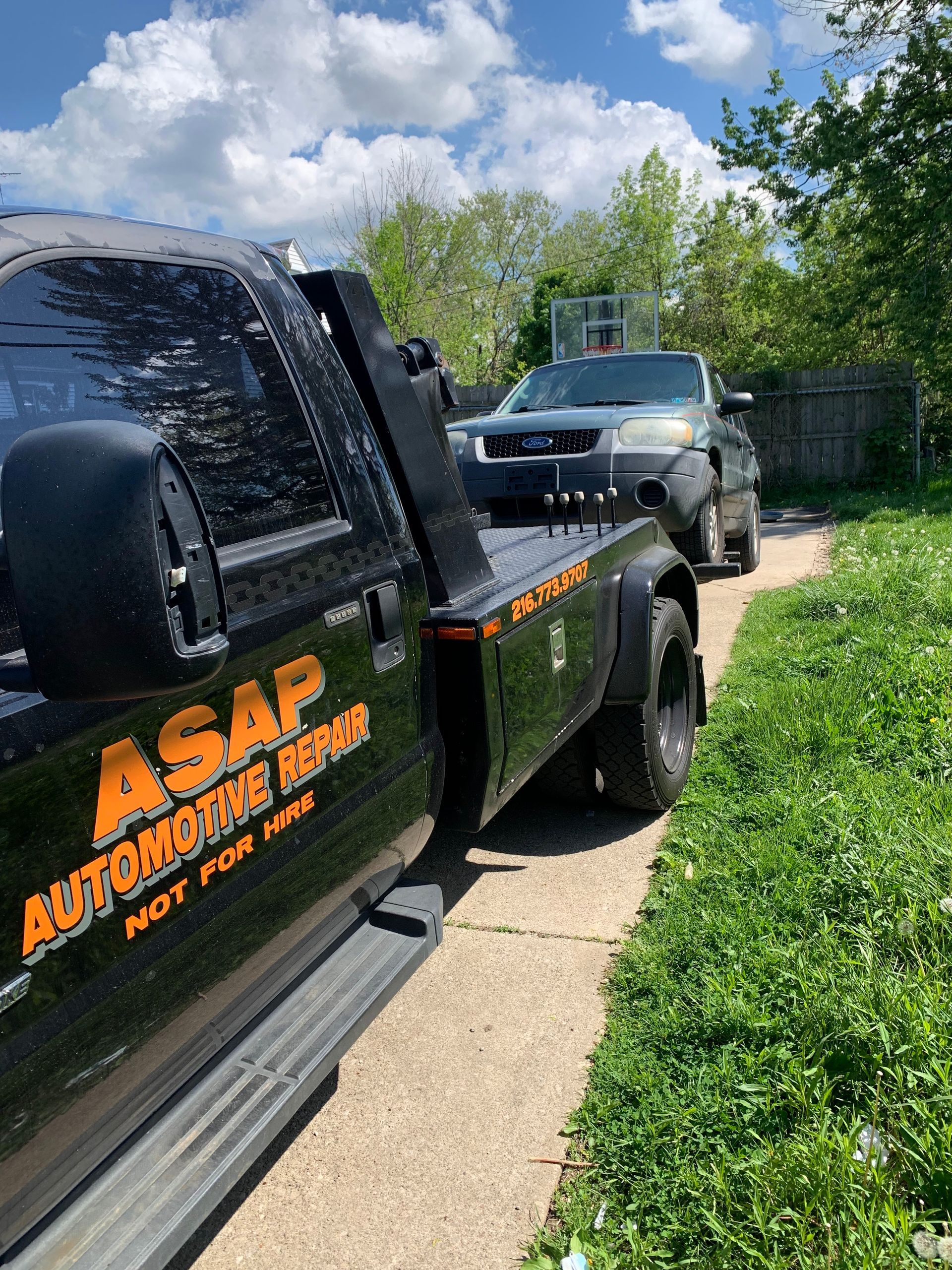 Black tow truck with orange logo towing a gray SUV along a grassy driveway under a blue sky