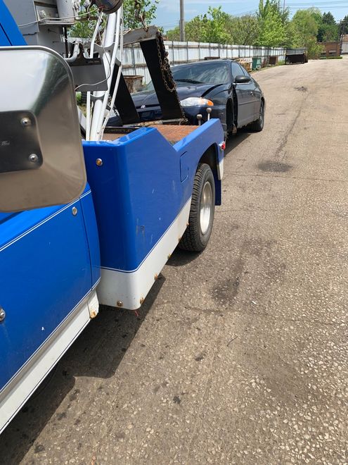 Blue tow truck hauling a black sedan along a gravel roadside under clear skies