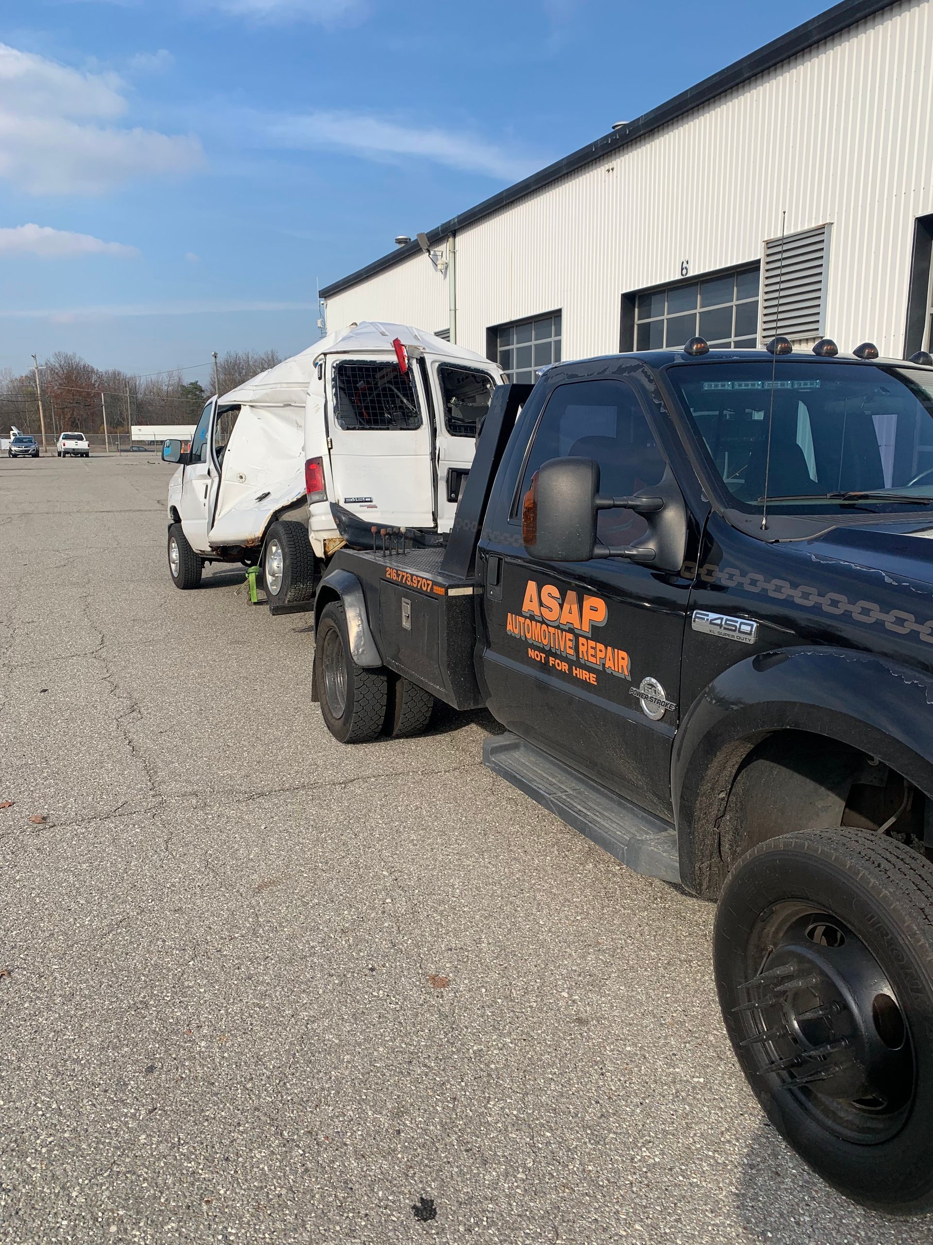 Tow truck hauling a damaged white van beside a warehouse on a gravel lot