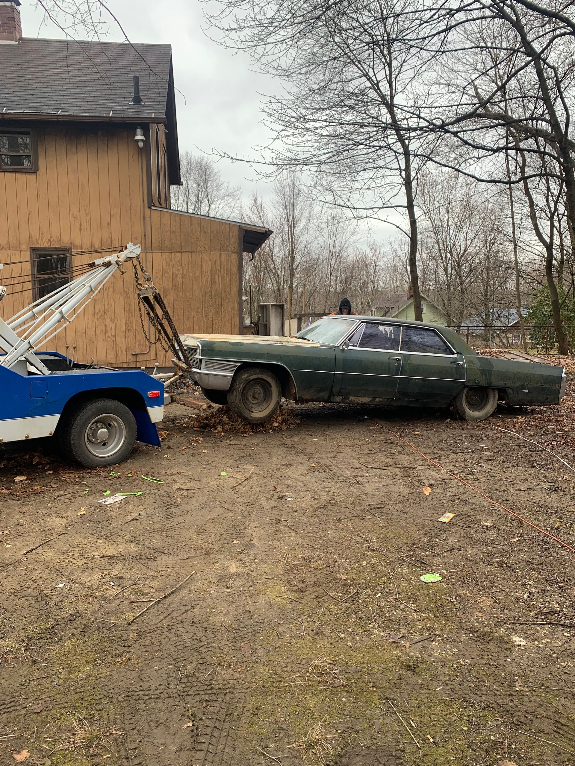 Green sedan being towed by a blue tow truck beside a house in a muddy yard.