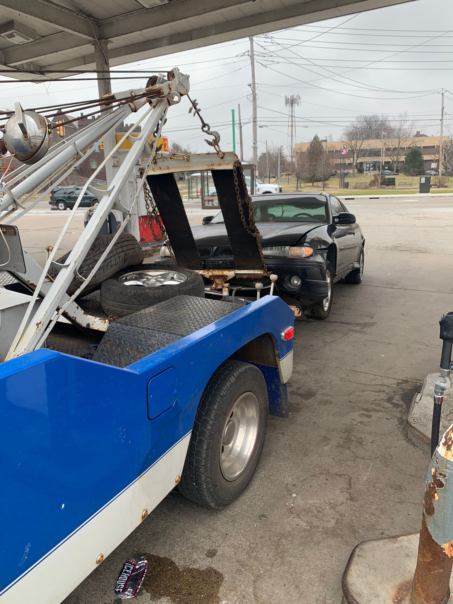 Tow truck under a gas station canopy, loading a black car in a parking lot.