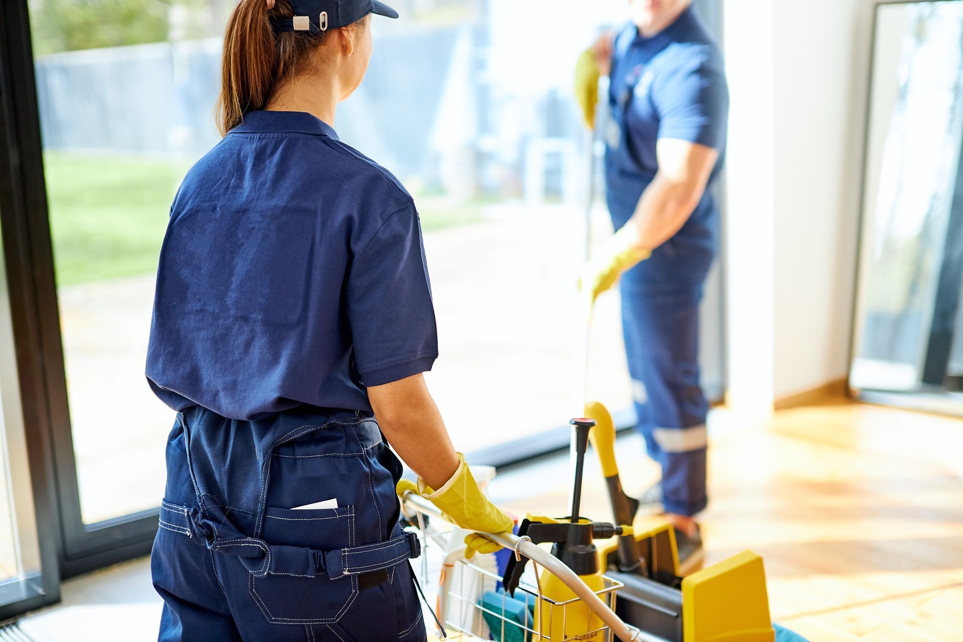 Reinigungskraft in blauer Uniform und gelben Handschuhen schiebt einen Reinigungswagen in einem hellen Raum