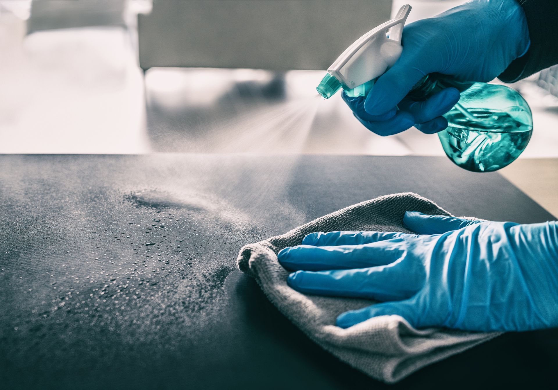 Mit blau behandschuhten Händen wird Desinfektionsmittel aufgesprüht und eine Arbeitsfläche mit einem Tuch abgewischt.