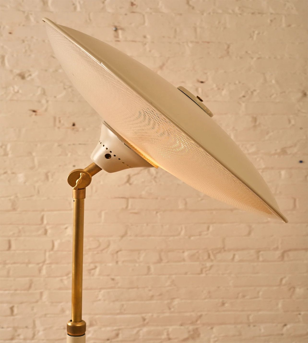 White, dish-shaped floor lamp on a brass pole, against a white brick wall.