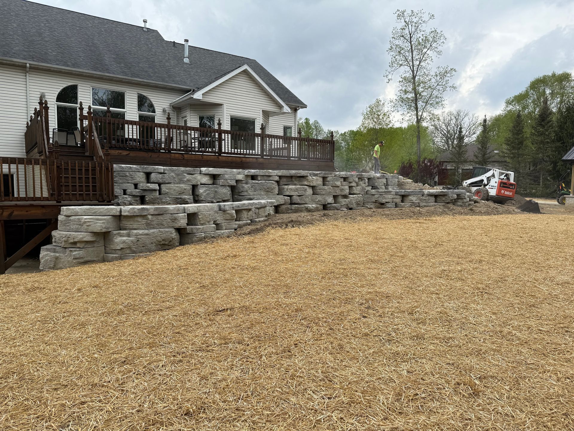 Stone retaining wall next to a wooden deck of a house on a cloudy day with a small excavator.