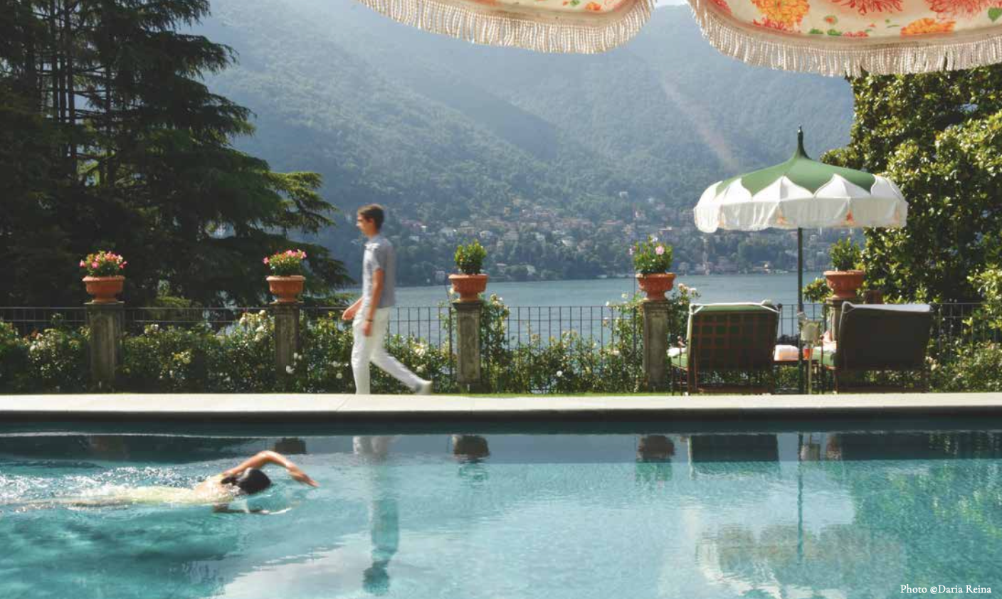 Man swims in pool, another walks by. Lake and mountains in the background. Green and white umbrellas overhead.