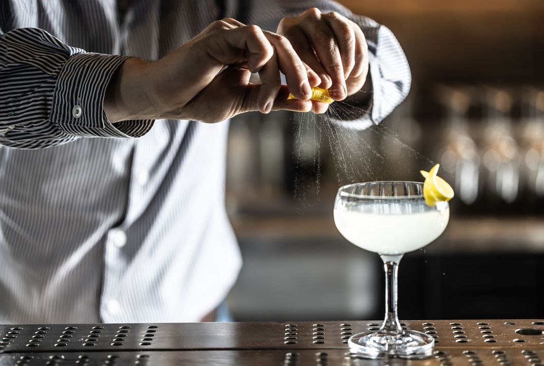 Bartender squeezing lemon zest over a cocktail in a coupe glass.