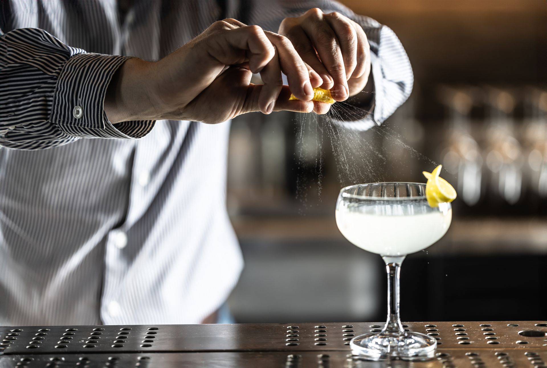 Bartender squeezing lemon zest over a cocktail in a coupe glass.