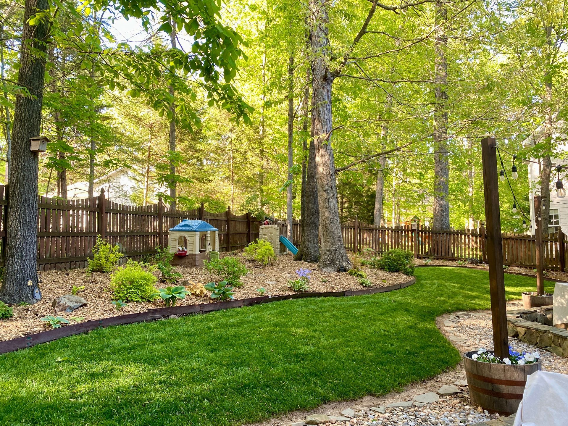 Leafy backyard with green lawn, tall trees, wooden fence, and a patio umbrella near a stone border