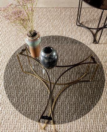 Un tavolino da caffè circolare in vetro con base in metallo dorato poggia su un tappeto dalla texture neutra, sul quale sono appoggiati due vasi.