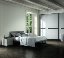 Camera da letto moderna con pareti bianche, letto, comodino, ampi armadi e pavimento scuro a spina di pesce vicino a una finestra.