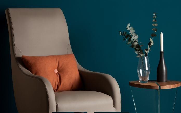 Una poltrona beige con un cuscino decorativo arancione è posizionata accanto a un tavolino su cui si trovano una candela e un vaso di piante.