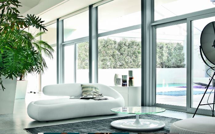 Soggiorno moderno con divano bianco dalle linee curve, tavolino da caffè in vetro, grande pianta da pavimento e finestre con vista sulla piscina.