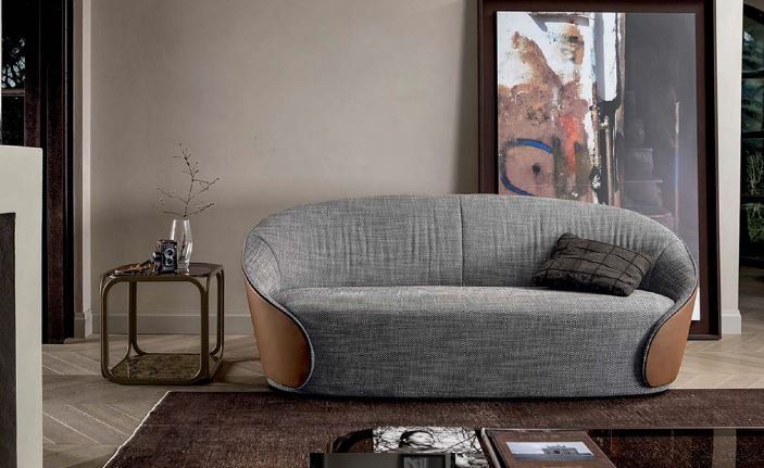 Un divano curvo in tessuto grigio con inserti in pelle color cuoio alle estremità, collocato in un soggiorno con un tavolino e una stampa artistica.