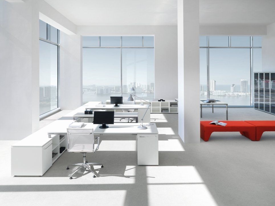 Un ufficio luminoso e moderno a pianta aperta con scrivanie bianche, arredi minimalisti, ampie finestre con vista sulla città e una panca rossa.