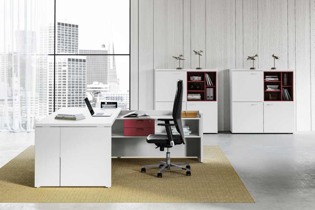 In un luminoso ufficio con vista sulla città, una scrivania e una sedia moderne di colore bianco con dettagli rossi, posizionate su un tappeto dalla texture particolare.