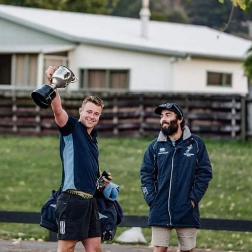 A smiling person holds a trophy aloft while standing beside a bearded person in a jacket on a field near a house.