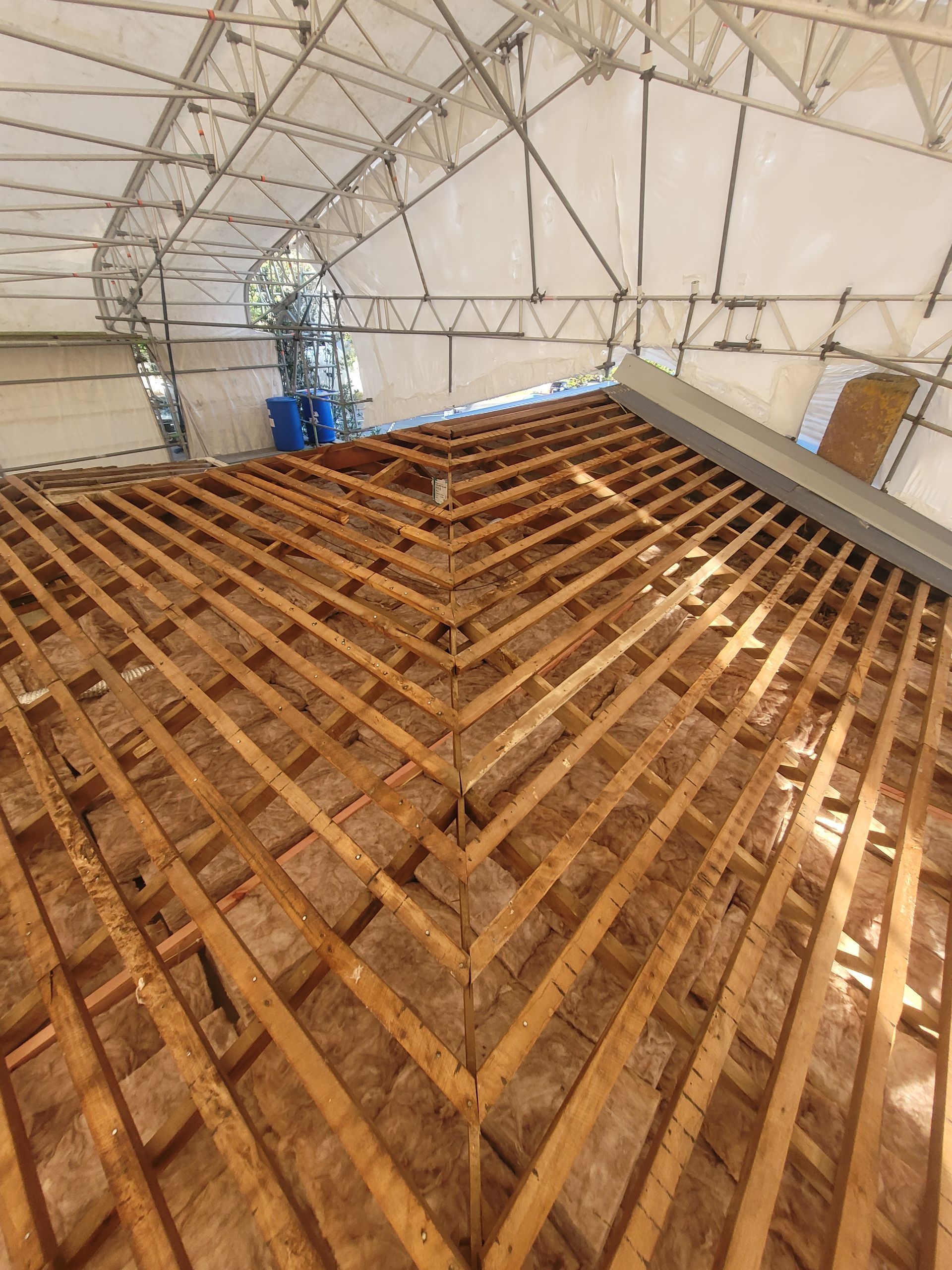 A high-angle view of a wooden roof frame under construction, inside a large, tented temporary shelter structure.