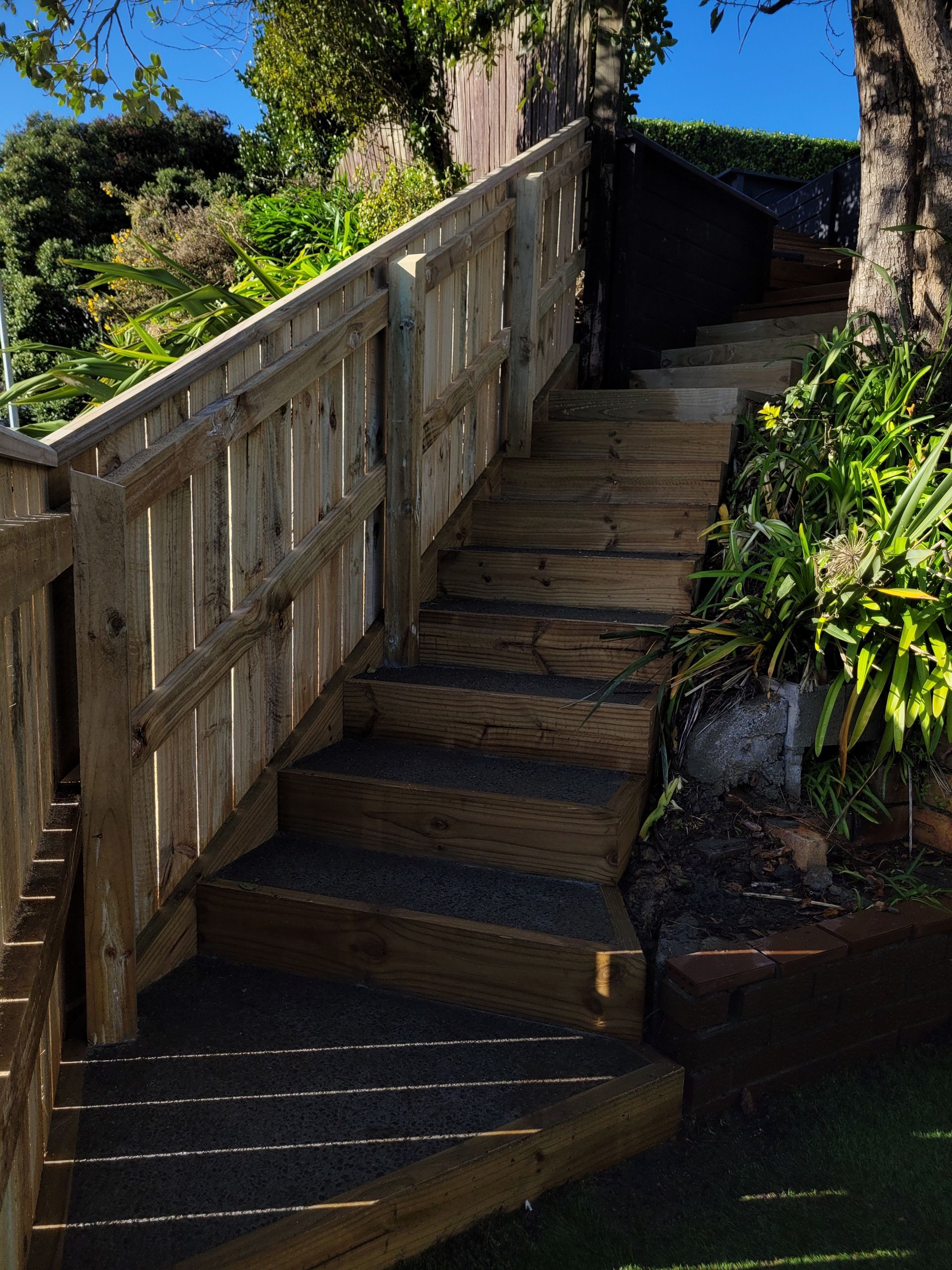 Outdoor wooden stairs with gravel treads climb a steep incline beside a wooden fence and lush greenery.
