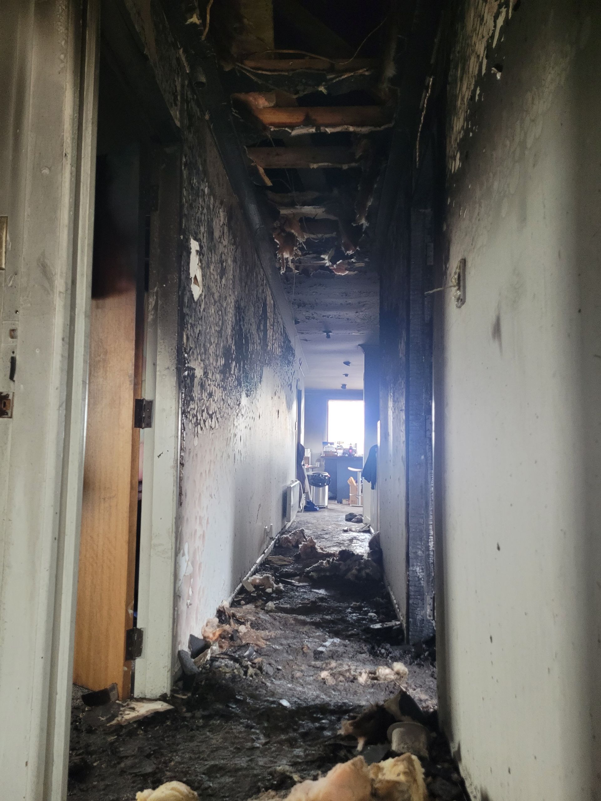 A narrow indoor hallway severely damaged by fire, with charred walls, a collapsed ceiling, and debris on the floor.