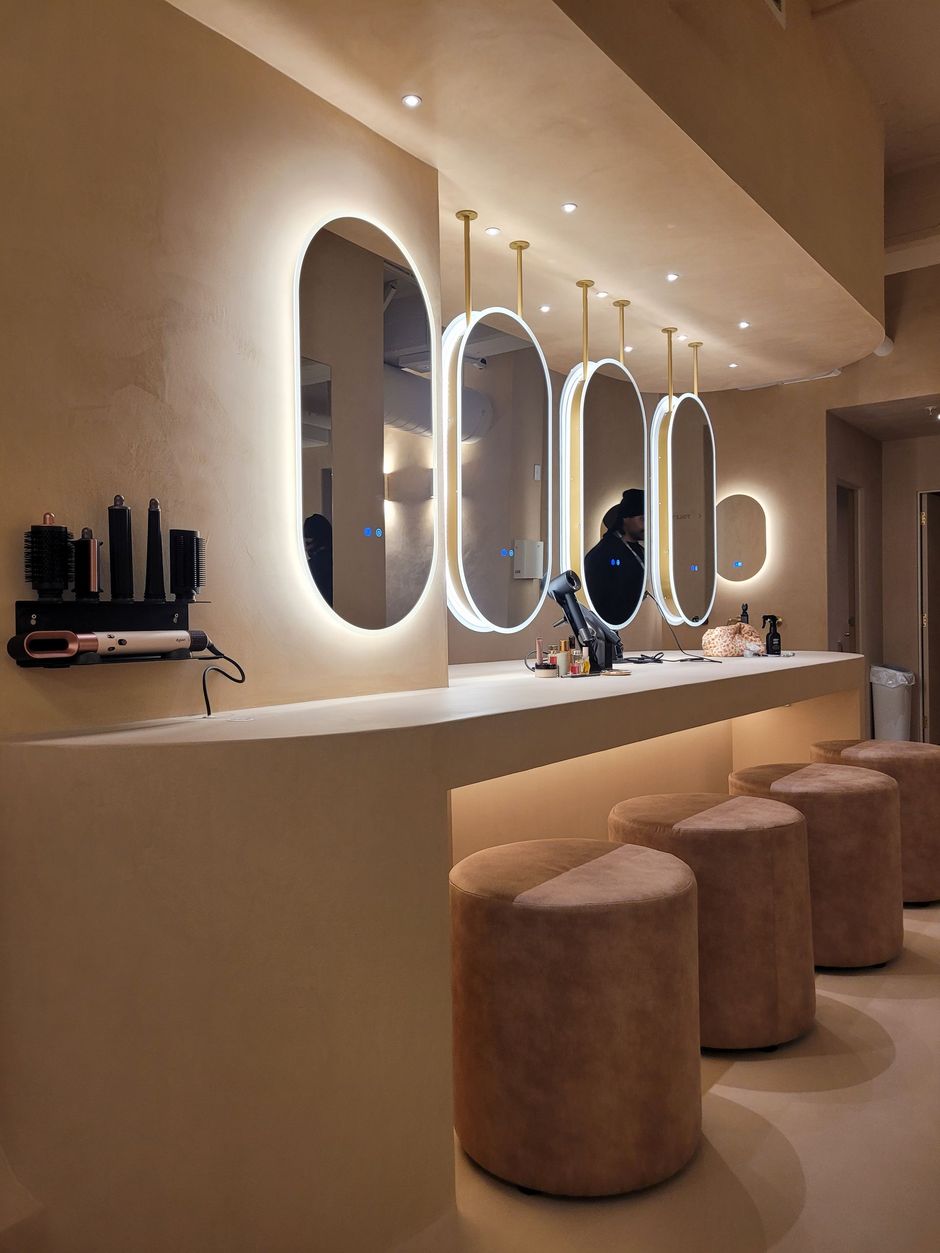 A modern salon featuring four backlit oval mirrors, a smooth beige counter, and matching round, velvet-textured stools.