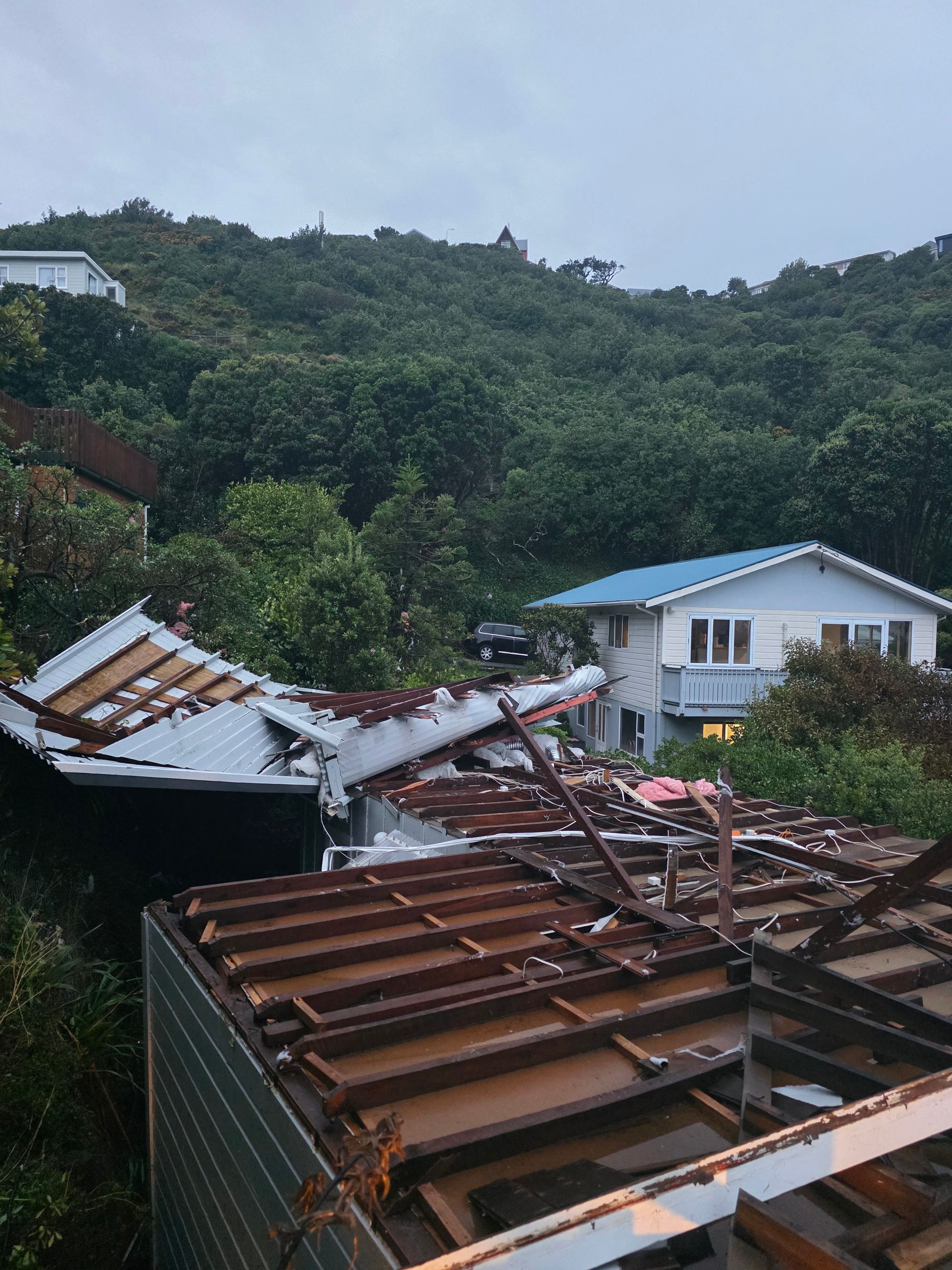 A house with a severely damaged, partially collapsed roof in a lush, green, hilly area.