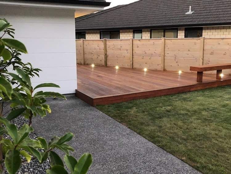 A wooden deck with built-in floor lighting, adjacent to a house, concrete path, and a lawn with a wooden fence.