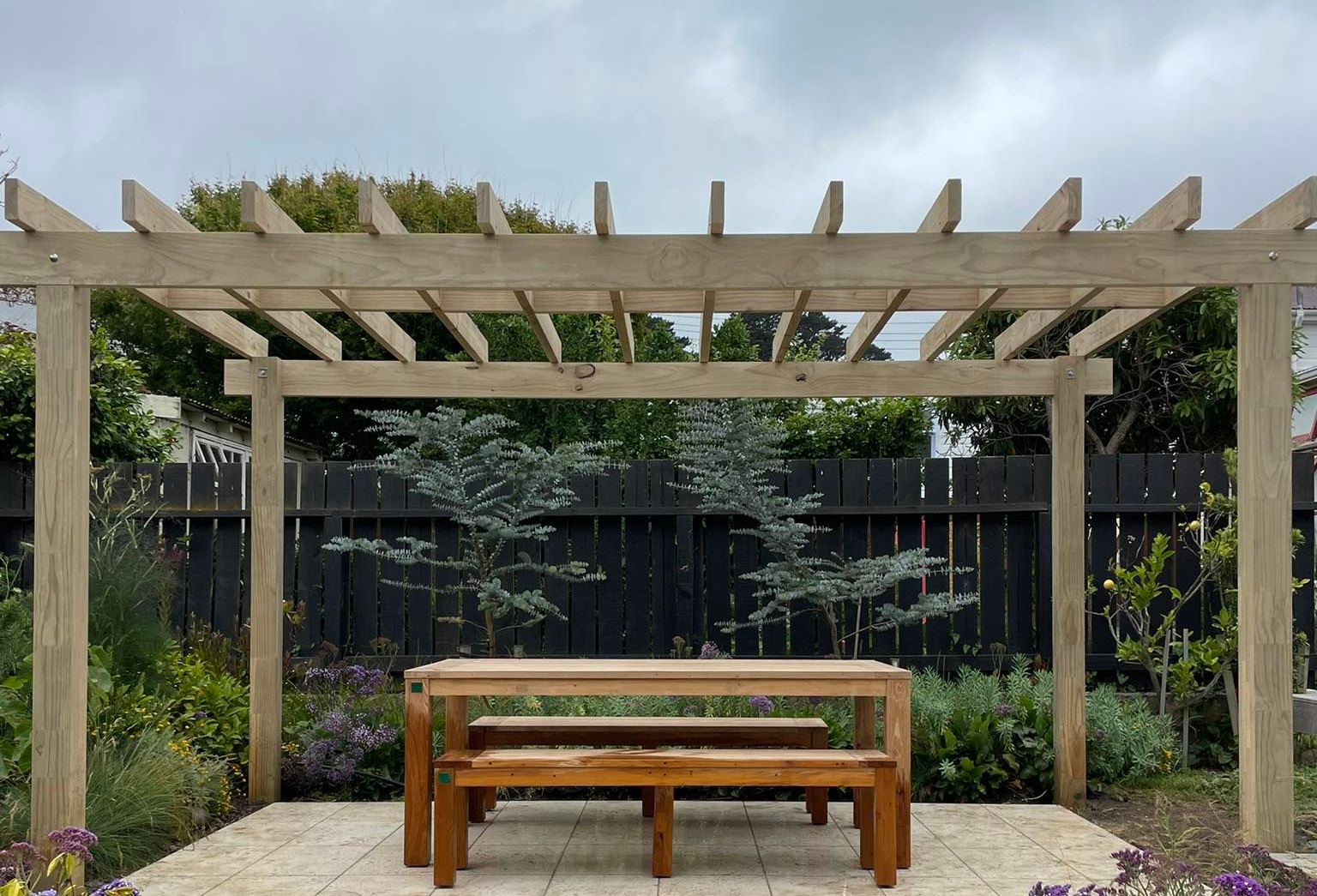 Wooden pergola over a dining set with a table and two benches on a light stone patio in a lush backyard.
