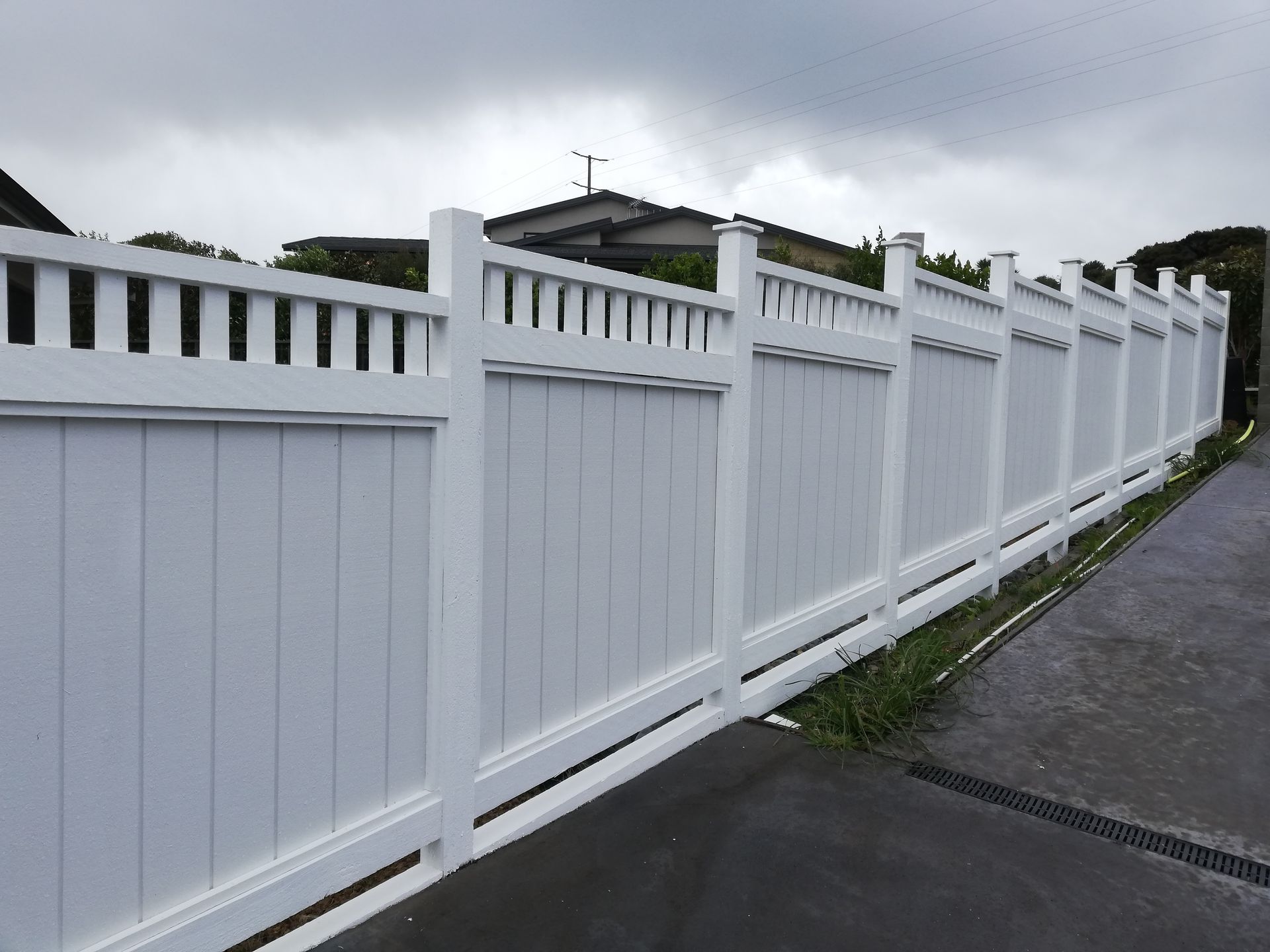 A white vinyl privacy fence with a lattice top border runs along a grey sidewalk under a cloudy sky.