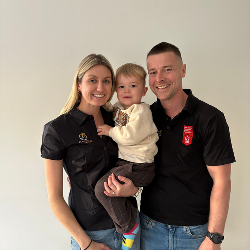 A smiling family of three posing for a photo against a plain, light-colored wall.