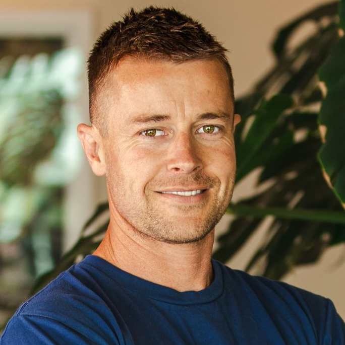 A person with short hair and a neutral expression wears a dark blue shirt against a background of green leaves.