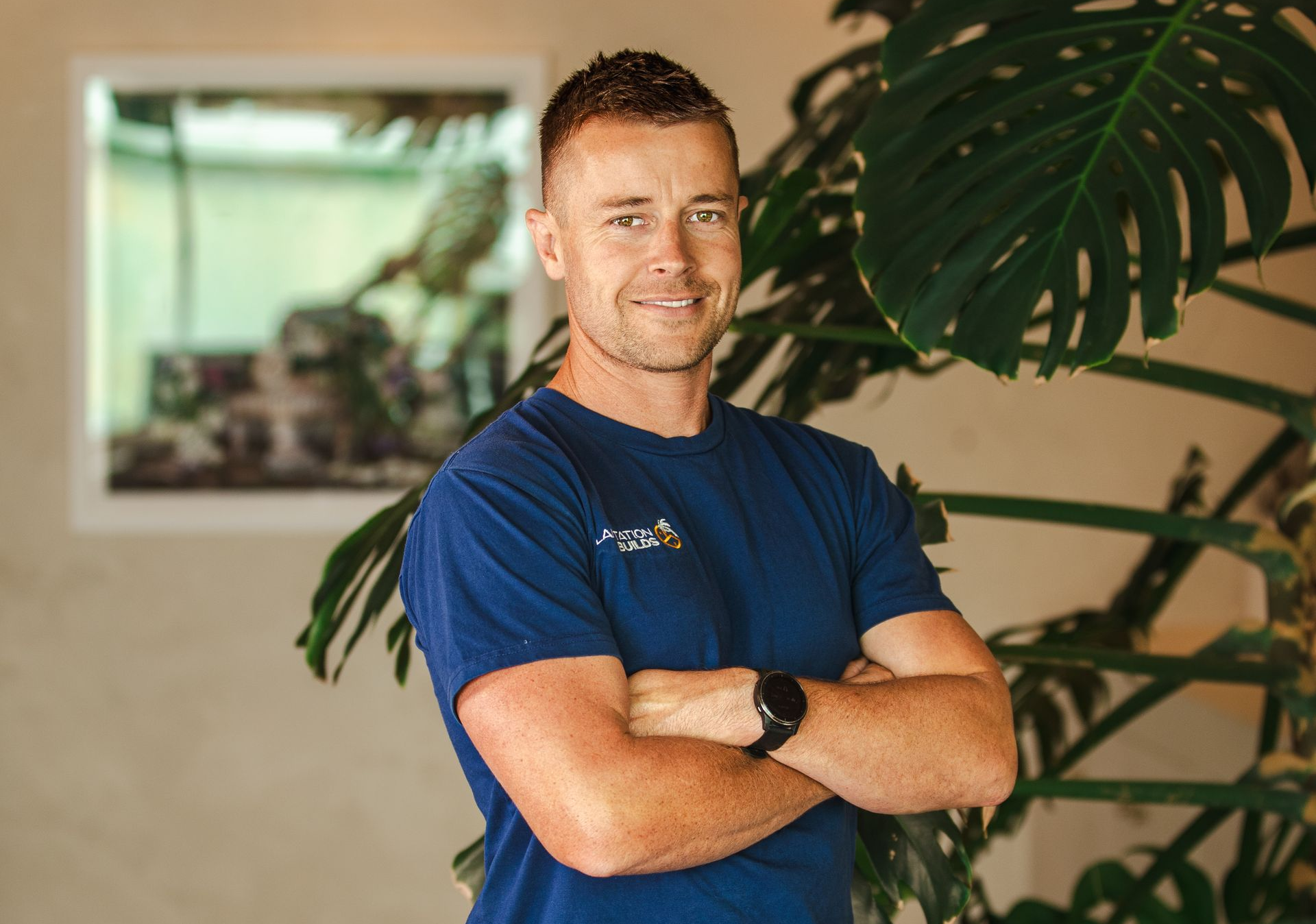 A person in a blue t-shirt with arms crossed stands smiling in front of a large, green monstera plant.