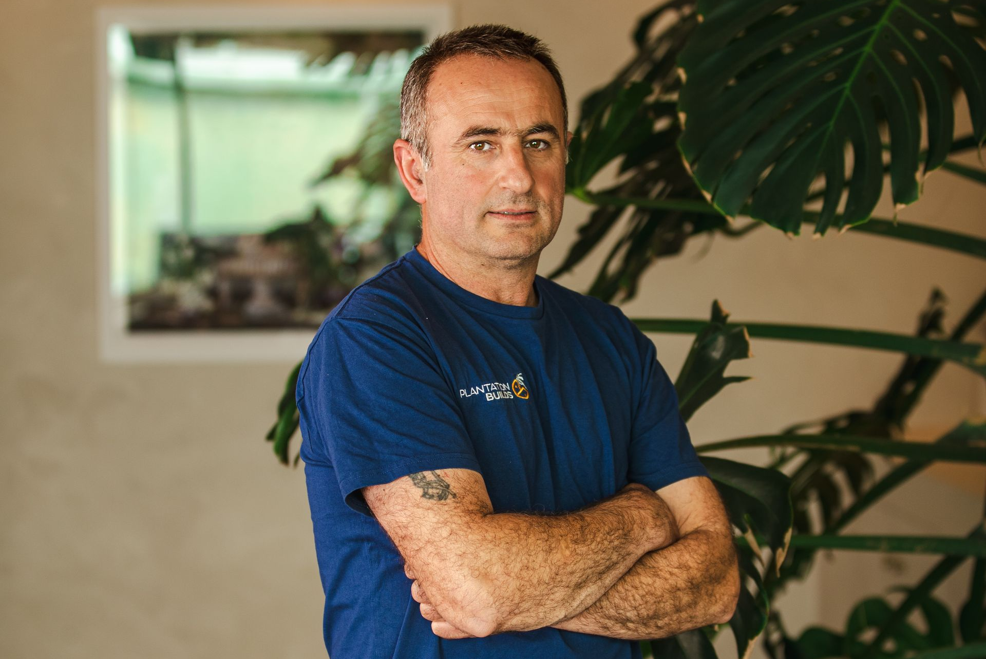 A person with a tattoo on their arm, wearing a blue t-shirt, stands with arms crossed before a large, leafy plant.