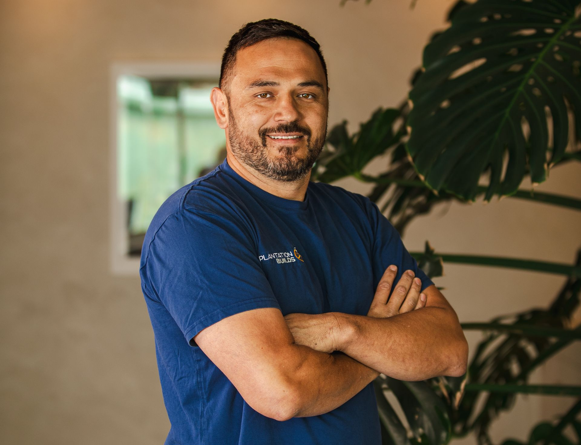 A smiling person with a beard, wearing a blue t-shirt with arms crossed, stands next to a large-leafed houseplant.