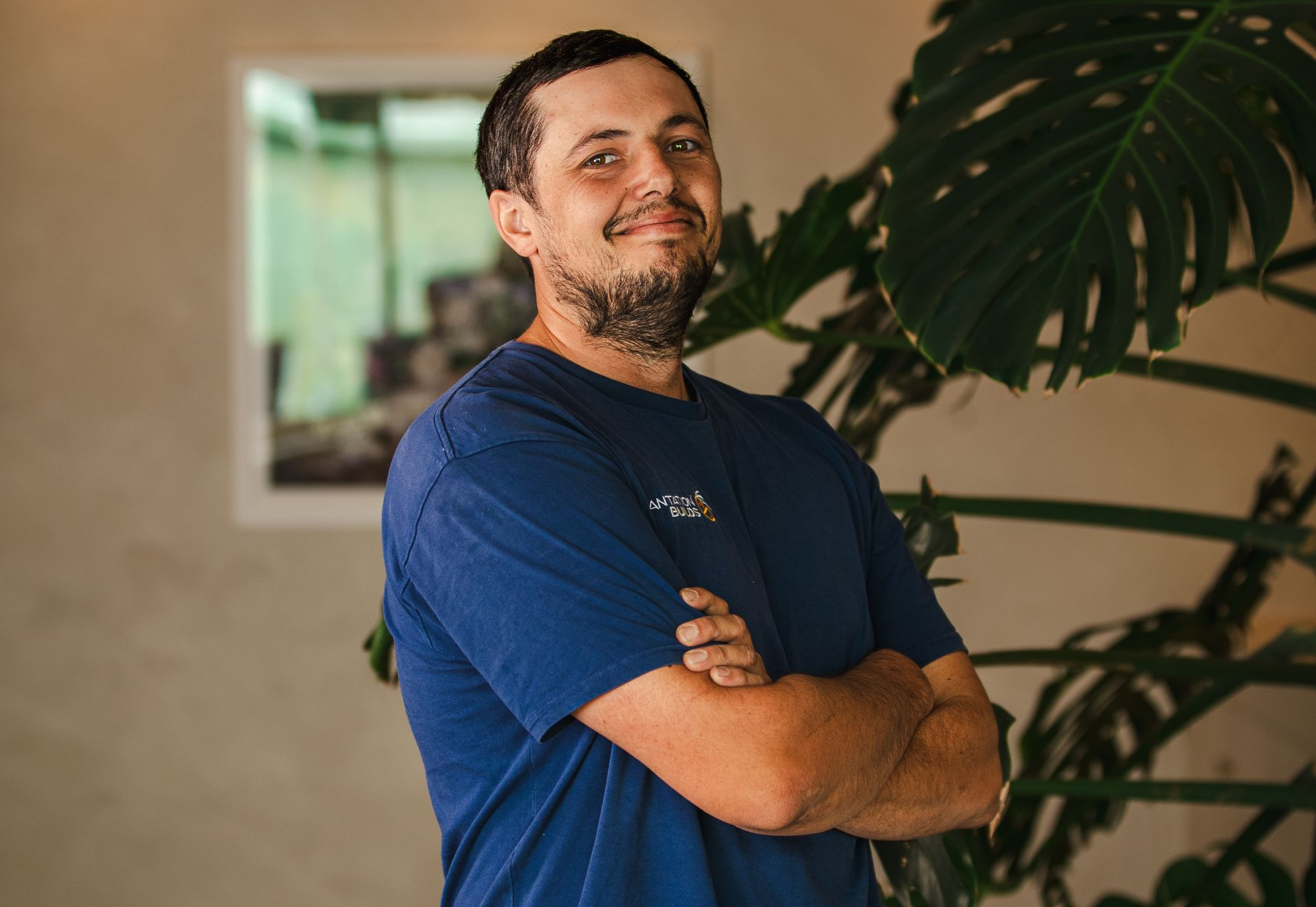 A person wearing a blue t-shirt stands with arms crossed, smiling, in front of a large indoor plant.