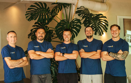 Five people in matching dark blue shirts stand with arms crossed before a large indoor plant.