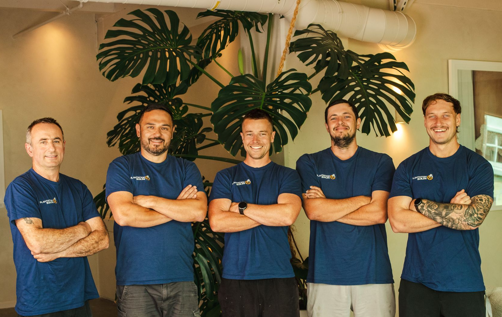 Five people stand in a row before a large potted plant, wearing identical navy blue shirts with their arms crossed.