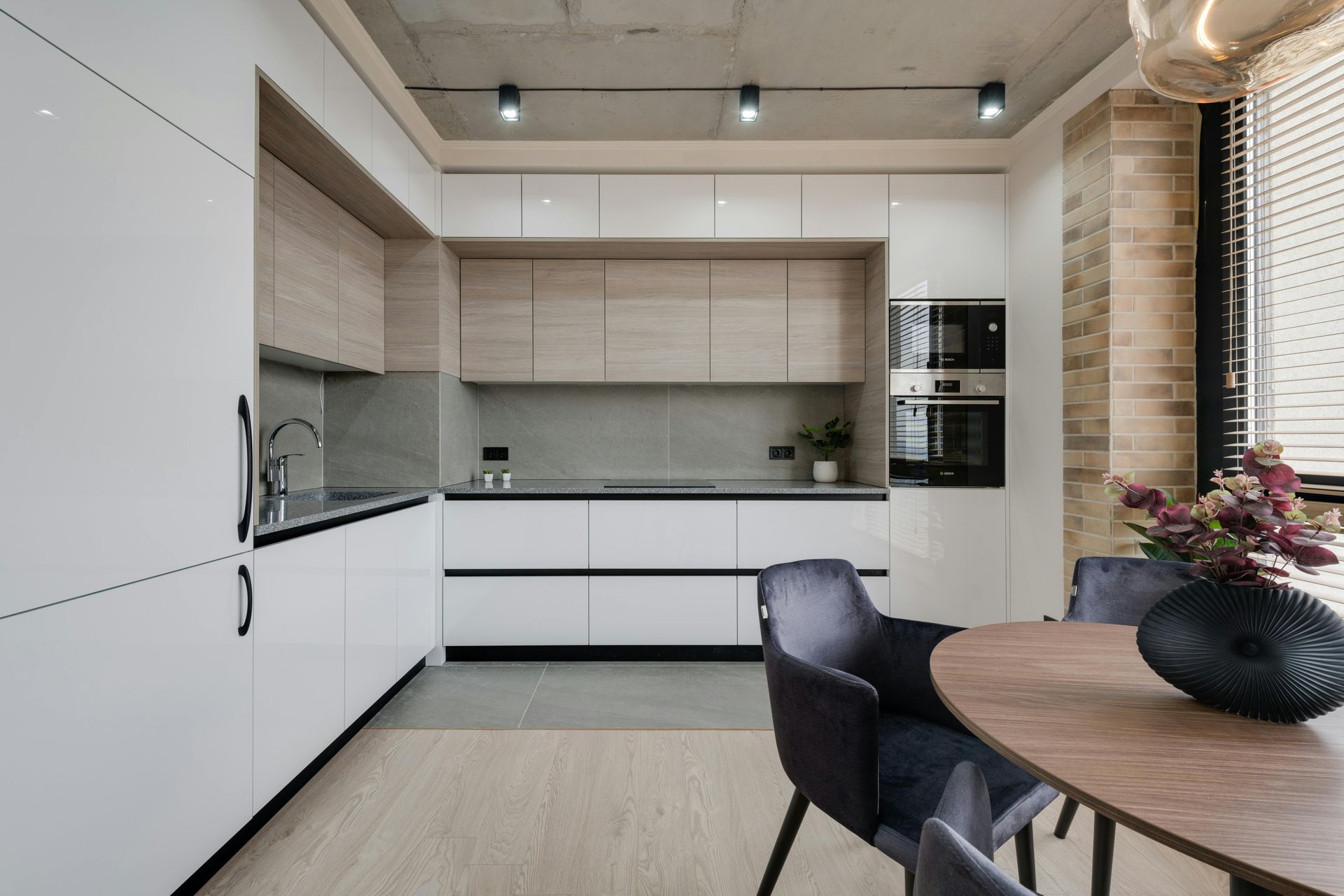 A modern kitchen with white cabinets, light wood accents, gray countertops, a concrete ceiling, and a round dining table.