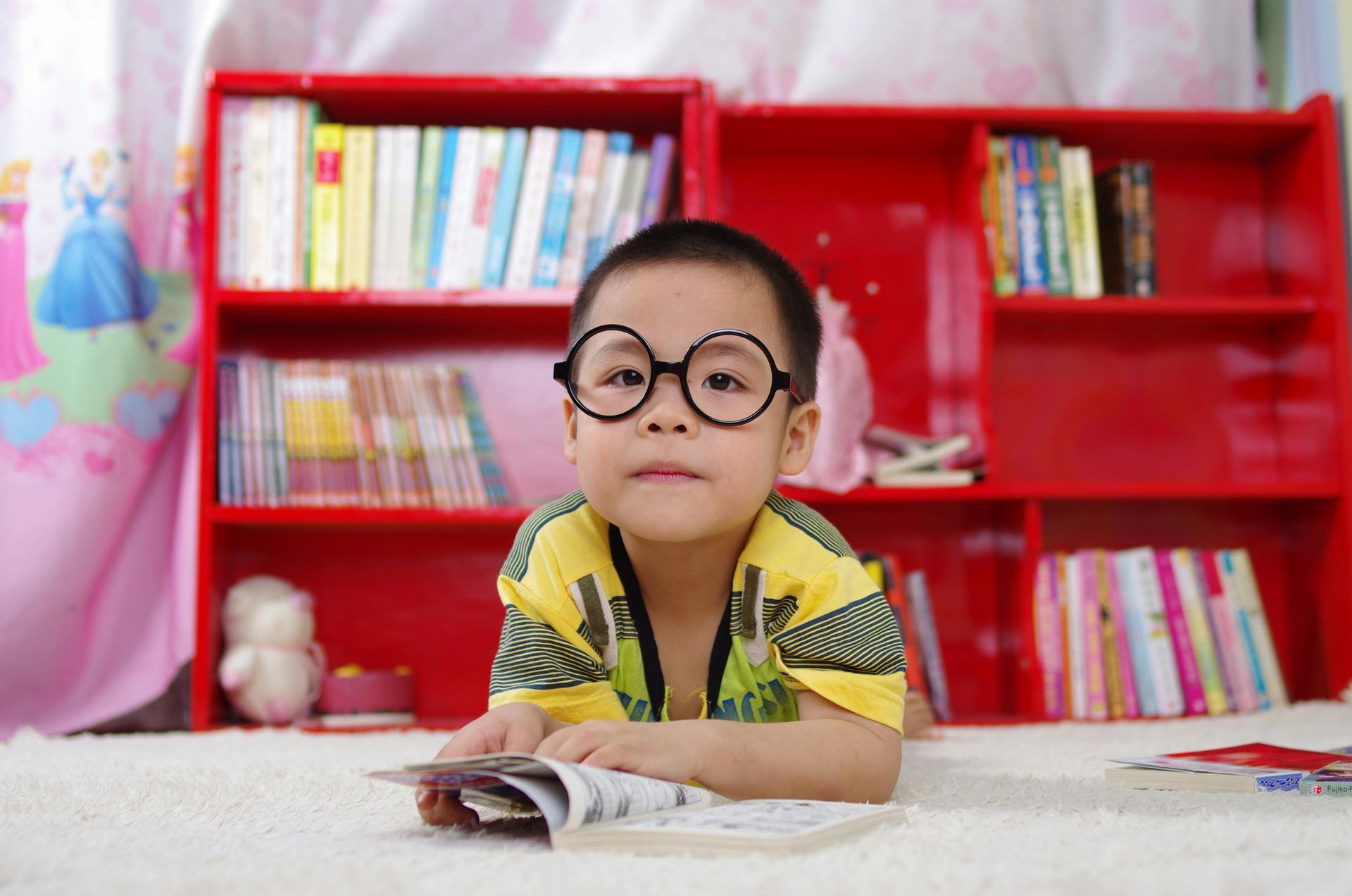 Young child with glasses reading a book in front of a red bookshelf.